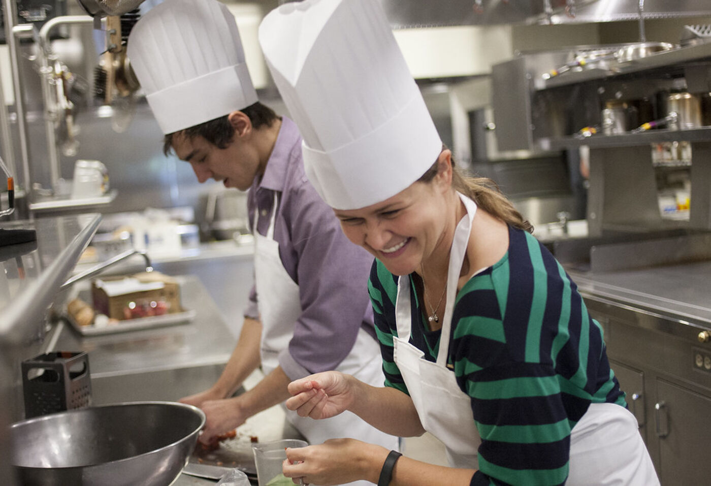 Two cooks in white aprons and chef hats laughing while they work